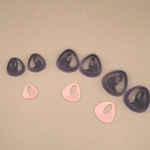 Può includere: Una collezione di sei tagliapasta in plastica viola e tre forme in argilla rosa chiaro. I tagliapasta hanno un contorno triangolare arrotondato irregolare con creste curve interne. Le tre forme rosa chiaro presentano un contorno irregolare abbinato con una serie di linee curve radianti in rilievo su un lato e un ritaglio circolare vicino alla parte superiore.