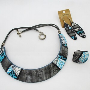 Puede incluir: Un conjunto de joyas que incluye un collar llamativo, pendientes y un anillo. El collar es una pieza ancha y curva hecha de arcilla polimérica gris y azul con un patrón geométrico. Los pendientes son largos y de forma ovalada, también hechos de arcilla polimérica gris y azul con un patrón geométrico. El anillo es de forma cuadrada con un patrón geométrico similar. Las joyas están hechas por Caristrass.