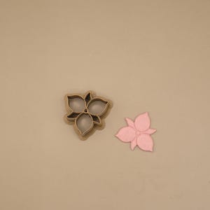 Puede incluir: Un cortador de galletas en forma de flor de tres pétalos de color beige con un orificio circular central, colocado junto a un recorte de flor de color rosa claro de tres pétalos que coincide con la forma del diseño interior del cortador.