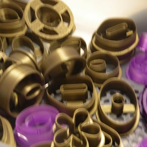 Pode incluir: Uma coleção de cortadores de biscoitos de plástico em dourado metálico e roxo translúcido com várias formas geométricas e florais. Os cortadores apresentam paredes finas e elevadas, dispostos num monte desordenado com algum foco.