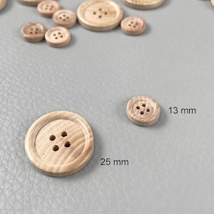Pode incluir: Uma coleção de botões de madeira castanha clara em vários tamanhos. O botão maior tem 25 mm de diâmetro, e o menor, 13 mm. Cada botão tem quatro orifícios para costurar e uma borda ligeiramente elevada.