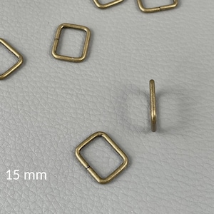 Pode incluir: Vários anéis quadrados em latão antigo, cada um com 15 mm. Um anel está posicionado de lado, mostrando sua espessura. Os anéis estão espalhados sobre uma superfície cinza, proporcionando um fundo neutro para o hardware.
