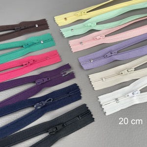 Pode incluir: Uma coleção de zíperes coloridos em várias tonalidades, incluindo marrom, verde, rosa, roxo e branco. Cada zíper tem aproximadamente 20 cm de comprimento e possui um cursor e dentes de metal. Os zíperes estão dispostos sobre uma superfície cinza.