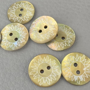 Può includere: Una collezione di bottoni rotondi e iridescenti con un disegno a forma di sole. Ogni bottone ha due fori e un motivo a raggiera bianco. I bottoni hanno una tonalità perlacea e dorata.