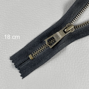 Può includere: Una cerniera nera, lunga 18 cm, con un tirante e denti in metallo color ottone. La cerniera è aperta, mostrando i dentini e il cursore. Il testo "18 cm" è visibile.