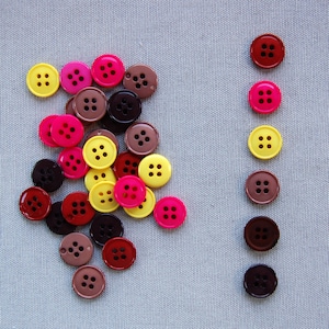 Puede incluir: Una colección de botones coloridos en tonos de rosa, amarillo, marrón y rojo, dispuestos sobre una tela azul claro. Los botones, cada uno con cuatro agujeros, están dispersos al azar, algunos alineados en una fila vertical.