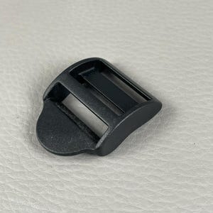 Pode incluir: Fivela tri-glide de plástico preto com um design curvo. A fivela tem duas ranhuras retangulares e uma aba arredondada. O objeto está sobre um fundo cinza claro.