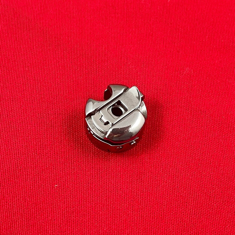 May include: A shiny, silver-colored sewing machine bobbin case. The bobbin case is made of metal and has a rounded shape with a central hole and a curved opening. The background is a solid red color.
