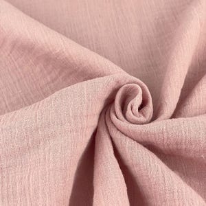Puede incluir: Primer plano de una tela rosa claro y suave con una apariencia texturizada y arrugada. La tela está doblada y drapeada, creando un patrón arremolinado. El material parece ligero y transpirable, adecuado para ropa o decoración del hogar.