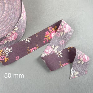 Pode incluir: Fita de viés de tecido floral, com 50 mm de largura, com um padrão repetido de flores rosa e amarelas sobre um fundo roxo escuro. A fita está desenrolada, mostrando o desenho floral. O texto "50 mm" é visível.