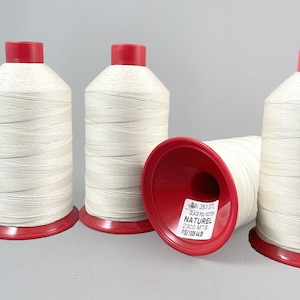 Puede incluir: Cuatro carretes de hilo blanco roto con tapas y bases rojas. Un carrete está tumbado de lado, revelando una etiqueta con texto. El hilo está bien enrollado, lo que sugiere un material de costura o artesanía de alta calidad.