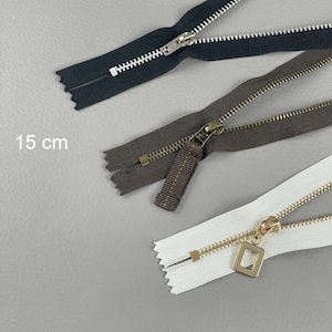 May include: Close-up of three zippers in black, brown, and white. The black zipper has a silver pull, the brown has a bronze pull, and the white has a gold pull with a square charm. The text "15 cm" is visible.