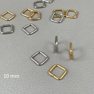 Può includere: Una collezione di anelli quadrati in metallo dorato e argentato. Gli anelli, di circa 10 mm, sono sparsi su una superficie grigia. Alcuni sono aperti, altri chiusi. Il testo "10 mm" è visibile.