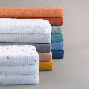 Pode incluir: Uma pilha de tecido dobrado e texturizado em várias cores, incluindo laranja, branco, cinza e azul. Algumas peças apresentam um padrão de estrelas. O tecido parece macio e leve, adequado para mantas de bebê ou artigos semelhantes.