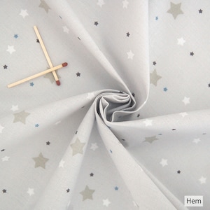 Pode incluir: Tecido cinza claro com um padrão repetido de estrelas brancas, cinzas e azuis. Dois fósforos de madeira estão colocados sobre o tecido. O tecido está dobrado, mostrando sua textura e design. O tema é celestial.