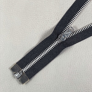 Pode incluir: Close-up de um fecho preto com dentes prateados e um puxador prateado. O fecho está aberto, revelando os dentes. O fecho está sobre uma superfície cinza claro.