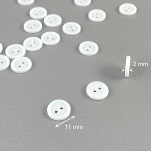 Pode incluir: Botões redondos brancos com dois furos, com cerca de 11 mm de diâmetro e 2 mm de espessura, espalhados sobre uma superfície cinza. Os botões são feitos de um material liso e sólido. A imagem mostra os botões de diferentes ângulos.