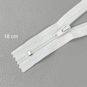Op de afbeelding: Een witte rits van 18 cm lang, met een witte schuif en tanden, ligt op een grijze ondergrond. De rits is open en toont de tanden. De meting "18 cm" is afgedrukt.