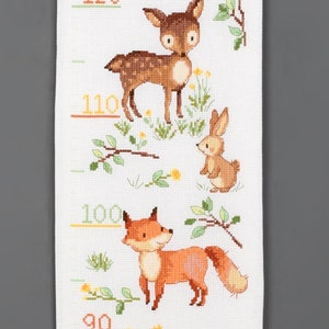 May include: A cross-stitched height chart featuring woodland animals. A brown deer, a tan rabbit, and an orange fox are depicted with yellow height markers. The chart is on a white background with green foliage and yellow flowers.