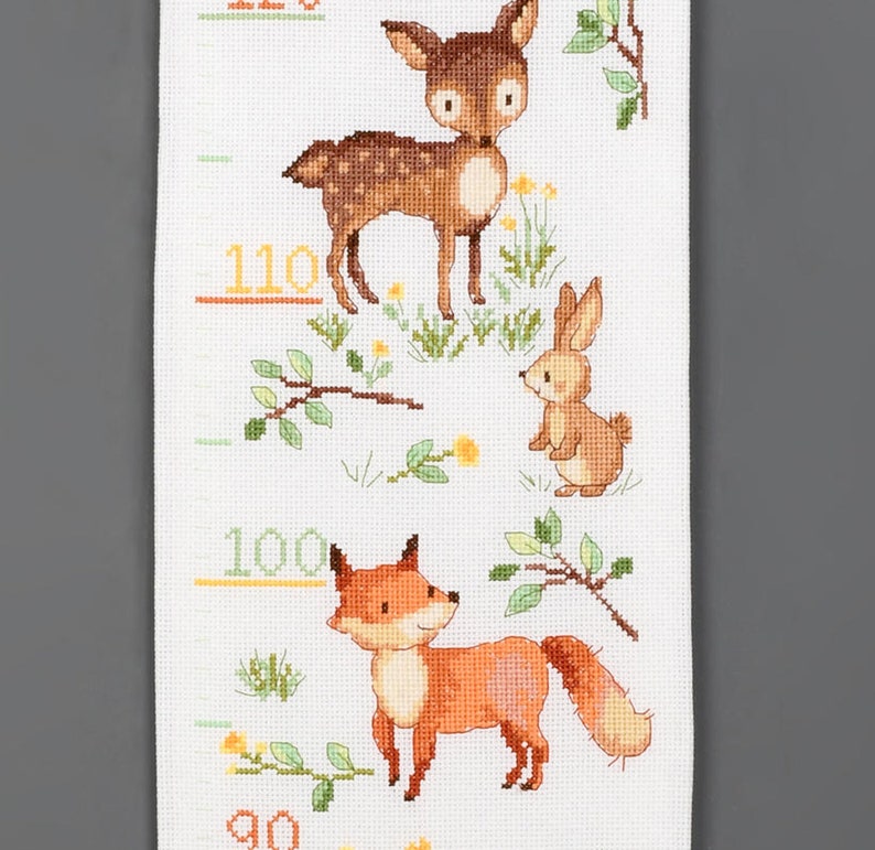 May include: A cross-stitched height chart featuring woodland animals. A brown deer, a tan rabbit, and an orange fox are depicted with yellow height markers. The chart is on a white background with green foliage and yellow flowers.