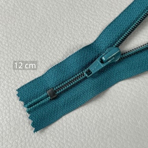 Pode incluir: Fecho de correr azul-petróleo, com aproximadamente 12 cm de comprimento, com um puxador a condizer. Os dentes do fecho são pretos e o tecido tem um aspeto texturizado. A imagem mostra o fecho de correr parcialmente aberto.