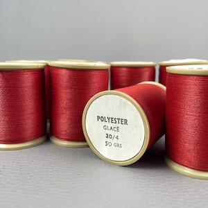 May include: Several spools of red polyester thread are arranged on a gray surface. One spool is lying on its side, revealing a label with the text "POLYESTER GLACÉ 20/4 50 GRS". The spools have beige-colored ends.
