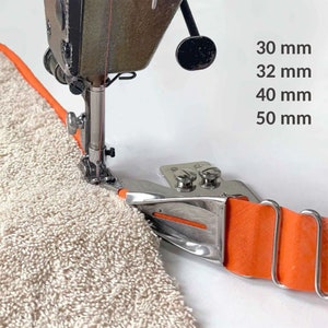 Peut inclure: Gros plan d'une machine à coudre avec un guide en métal fixée, guidant une bande de tissu orange le long du bord d'une serviette beige. Le guide affiche des mesures de 30 mm, 32 mm, 40 mm et 50 mm. La machine est en marche.