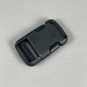 May include: A black plastic side-release buckle. The buckle has a rectangular frame with a series of teeth on one side and a snap-in closure on the other. The buckle is designed to secure straps or webbing.