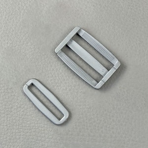 Pode incluir: Duas fivelas de metal prateadas. Uma é uma fivela deslizante retangular com três ranhuras paralelas em cada lado. A outra é uma fivela menor, em forma oval. Ambas estão sobre um fundo cinza texturizado.