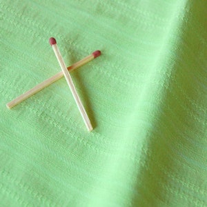 Pode incluir: Dois fósforos de madeira com cabeças vermelhas cruzam-se sobre um tecido verde claro texturizado. Os fósforos estão posicionados diagonalmente, formando um X sobre o fundo liso. O tecido tem um padrão subtil de riscas.