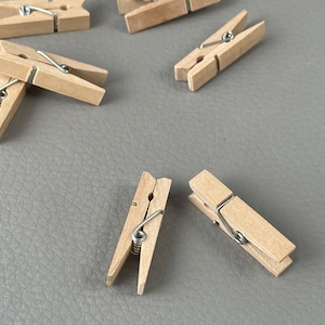 Può includere: Diverse piccole mollette di legno naturale con molle in metallo color argento sono sparse su una superficie grigia. Le mollette sono in legno chiaro e hanno un design classico.