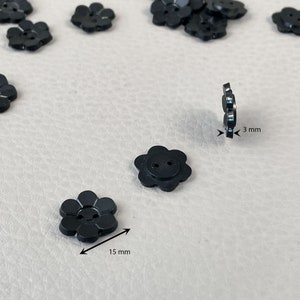 Pode incluir: Botões pretos em forma de flor, cada um com 1,5 cm de diâmetro e 3 mm de espessura. Os botões têm dois orifícios para costurar. Vários botões estão espalhados numa superfície clara.