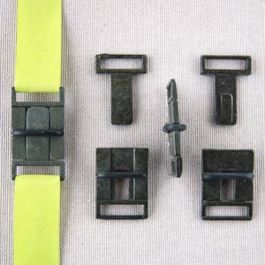May include: A collection of dark metal hardware components, including rectangular buckles and T-shaped connectors. A bright yellow fabric strap is attached to one of the buckles. The hardware has a weathered, antique appearance.
