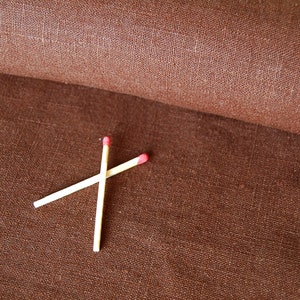 Pode incluir: Dois fósforos de madeira com pontas vermelhas cruzadas sobre uma superfície de tecido castanho texturizado. O tecido parece estar enrolado ao fundo, mostrando um tom mais escuro da mesma cor castanha. Os fósforos estão posicionados no centro inferior da imagem.