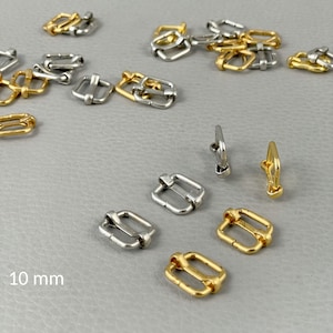 Pode incluir: Uma variedade de pequenas fivelas de metal em tons dourados e prateados. As fivelas, de forma retangular com uma barra central, medem aproximadamente 10 mm. Algumas têm uma argola para fixação.