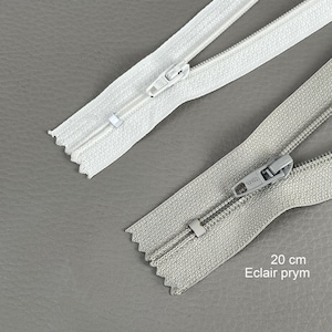 Pode incluir: Duas zíperes, uma branca e outra cinza, exibidas sobre um fundo cinza. A zíper cinza tem o texto "20 cm Eclair prym" impresso. Ambas as zíperes medem aproximadamente 20 cm de comprimento.
