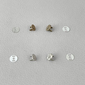 May include: A collection of magnetic snap closures in bronze and silver tones. Each closure consists of a round metal disc and a matching snap component. The components are arranged on a light gray surface.