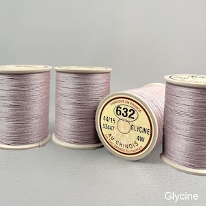 May include: Four spools of light purple sewing thread wound on beige plastic bobbins. The label on the front spool reads FABRIQUÉ EN FRANCE 632 44/15 53447 GLYCINE 4W AU CHINOIS ETS TOULEMONDE SUCCESSEURS. The thread has a smooth, slightly shiny finish.