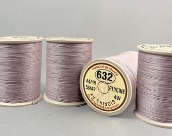 May include: Four spools of light purple sewing thread wound on beige plastic bobbins. The label on the front spool reads FABRIQUÉ EN FRANCE 632 44/15 53447 GLYCINE 4W AU CHINOIS ETS TOULEMONDE SUCCESSEURS. The thread has a smooth, slightly shiny finish.