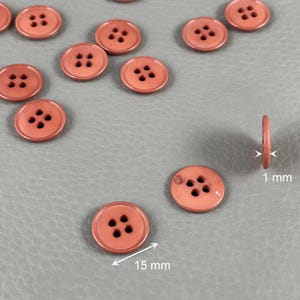 Può includere: Una collezione di bottoni rotondi color corallo con quattro fori. I bottoni hanno un diametro di 15 mm e uno spessore di 1 mm. I bottoni sono sparsi su una superficie grigia.