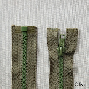 Pode incluir: Close-up de dois zíperes de cor azeitona. Um zíper está fechado e o outro está aberto, mostrando os dentes e o cursor. Os zíperes são feitos de plástico e têm um suporte de tecido texturizado. A palavra "Olive" é visível.