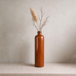 Peut inclure: Un vase en céramique marron, de forme élancée, contenant des herbes ornementales séchées et des brindilles. Le vase, au fini brillant, est posé sur une surface neutre, créant une esthétique minimaliste. Le vase mesure environ 30 cm de haut.