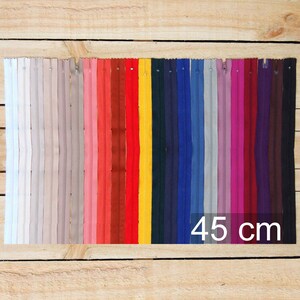May include: A collection of colorful zippers in various shades, including white, pink, red, yellow, blue, and purple. Each zipper is 45 cm (17.7 inches) long, displayed against a wooden background. The image showcases a range of colors for sewing and crafting projects.