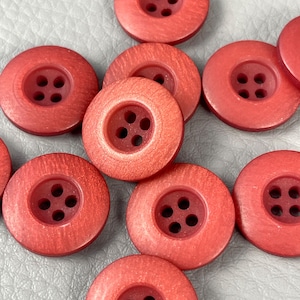 Può includere: Primo piano di diversi bottoni rotondi rossi. Ogni bottone ha un'area centrale incassata con quattro fori per cucire. I bottoni hanno una superficie leggermente strutturata e una tonalità rosso-arancio, che suggerisce un aspetto simile al legno.