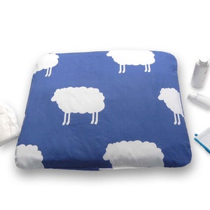 Peut inclure: Un matelas à langer bleu avec un motif de moutons blancs. Le matelas est rectangulaire avec des coins arrondis. D'autres articles de soins pour bébés sont visibles en arrière-plan.