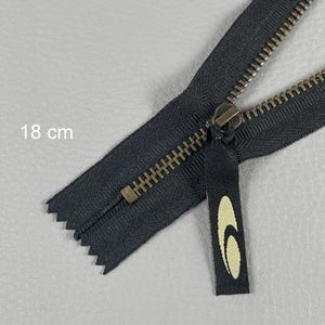 May include: A black zipper, approximately 7 inches long, with a brass-colored pull and teeth. The zipper pull features a black tag with a cream-colored logo. The image shows the zipper partially open.