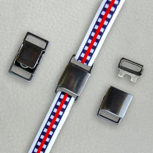 Pode incluir: Close-up de uma fita elástica branca com uma faixa central vermelha e um padrão xadrez azul. A fita é conectada com fechos e fivelas de metal prateado. Fechos e fivelas adicionais estão espalhados.