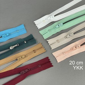 Pode incluir: Variedade de zíperes YKK de 20 cm, em cores como azul, verde, bege, rosa e bordô. Os zíperes são exibidos em uma superfície cinza.