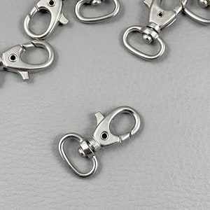 May include: Several silver-colored swivel snap hooks are scattered on a gray surface. The hooks have a spring-loaded clasp and a circular ring for attaching to other items. The metal has a shiny, reflective finish.