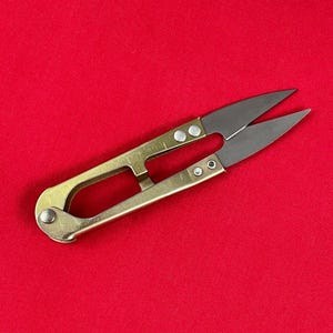 Peut inclure: Petites ciseaux à manche en laiton avec des lames argentées et tranchantes. Ces ciseaux sont conçus pour une coupe de précision, probablement pour la couture ou l'artisanat. Fond rouge uni.